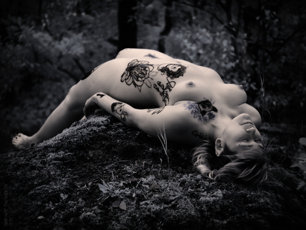 Tetované nahé ženské tělo zasazené do přírody. Černobílý snímek s jemným modrým tónováním.